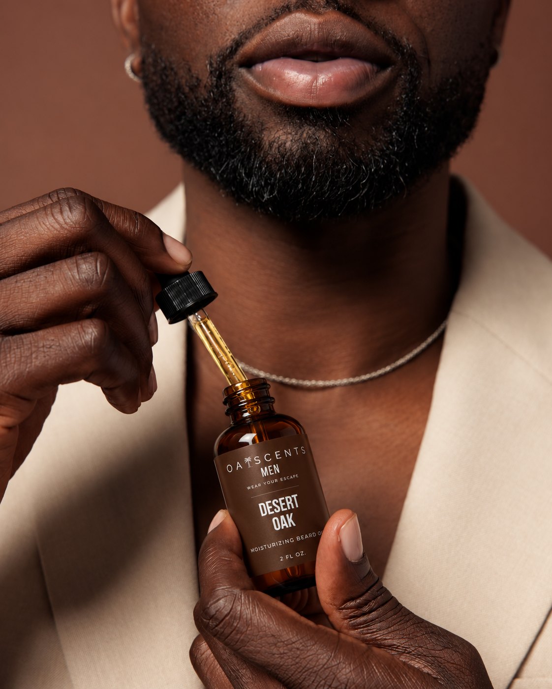 A man in a cream blazer holding a bottle of Oaiscents Desert Oak Moisturizing Beard Oil and lifting the glass dropper, against a warm terra-cotta backdrop