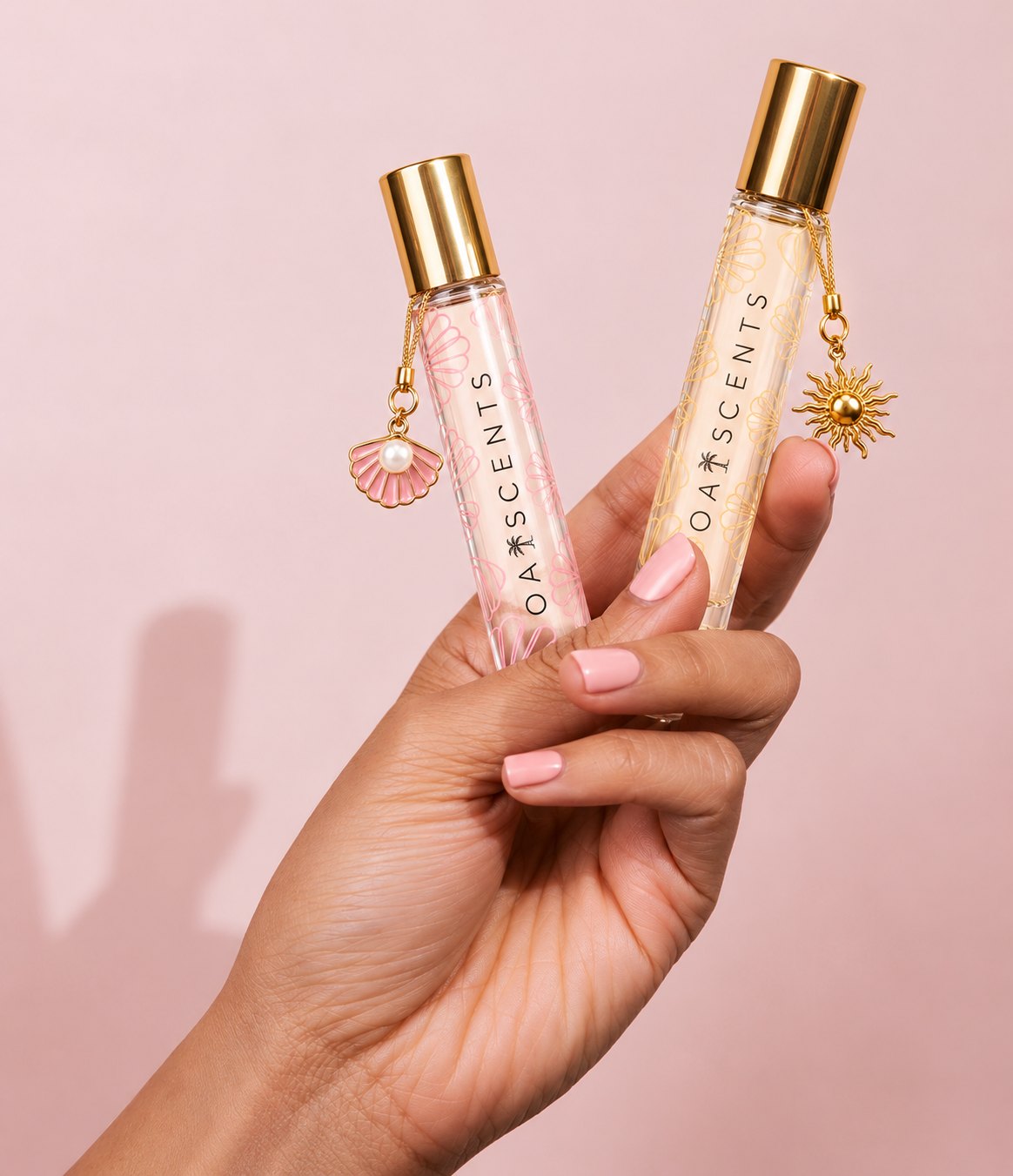 A hand holding two Oaiscents roll-on perfume oils — one with a pink seashell charm, one with a gold sun charm — against a soft blush backdrop