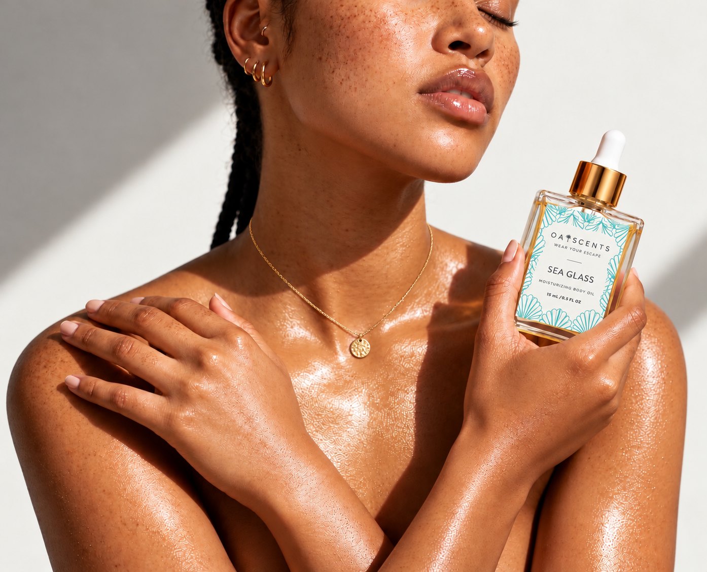 A woman with sun-kissed, oiled skin holding a bottle of Oaiscents Sea Glass Moisturizing Body Oil against a pale wall in warm sunlight